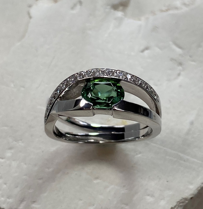 14 Karat White Gold ring with an oval Green Tourmaline in the center with split bands on one side and a crossover band with diamonds on the other.