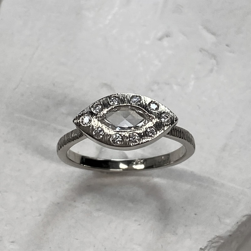 A white gold ring with a marquise shaped rose cut diamond with a halo around it with flush-set diamonds and a thin textured band.