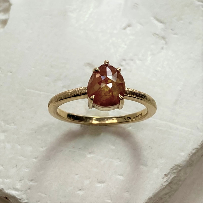Yellow gold ring with one orange-ish pear shaped diamond in a four prong setting and a thin textured band.