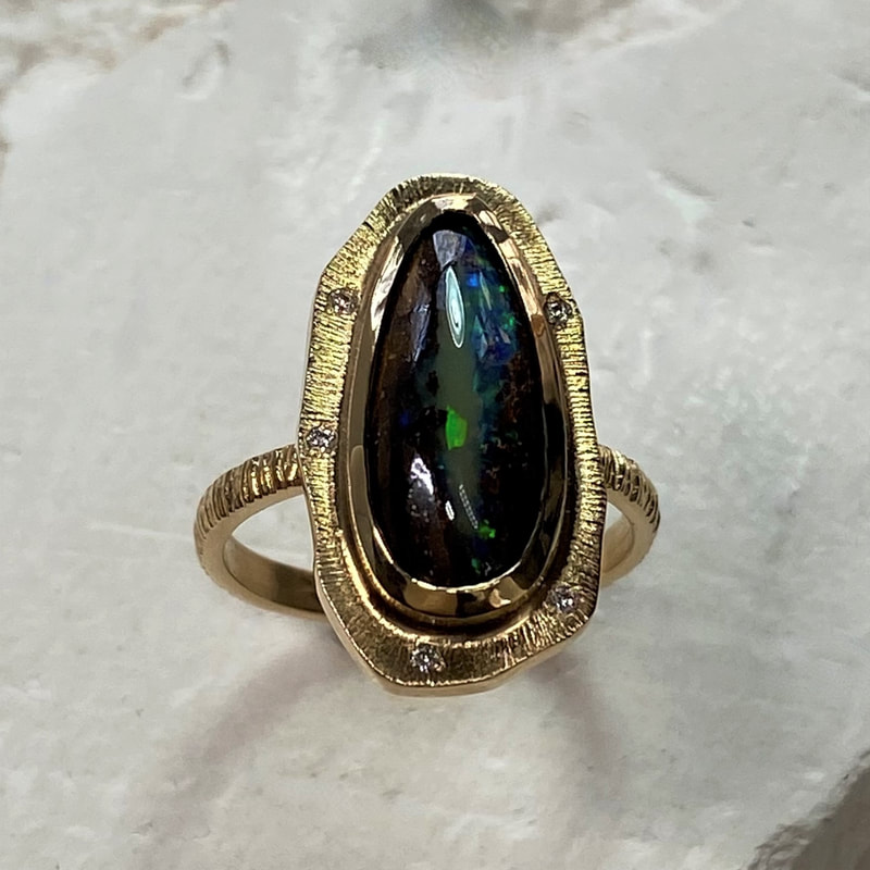 A gold ring with a natural elongated black opal and a border with spaced inset diamonds and a textured band.