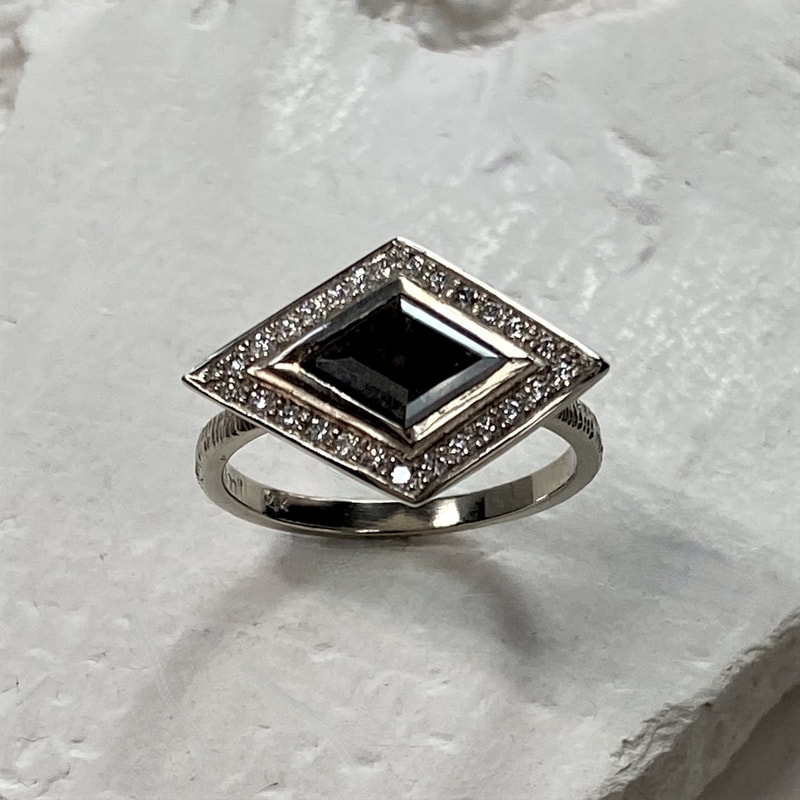 White gold ring with a black diamond shaped diamond in the center with white diamonds tracing the outside, set east to west.